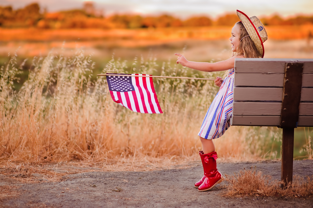 A young girl holding an American Flag and sitting on a bench