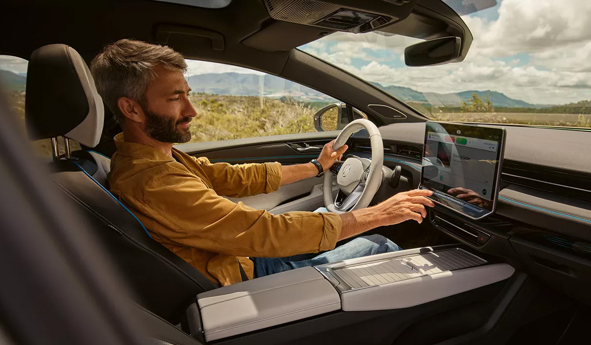 A bearded man in a yellow, collared shirt happily adjusting the settings in his Volkswagen vehicle while driving down the road on a sunny day